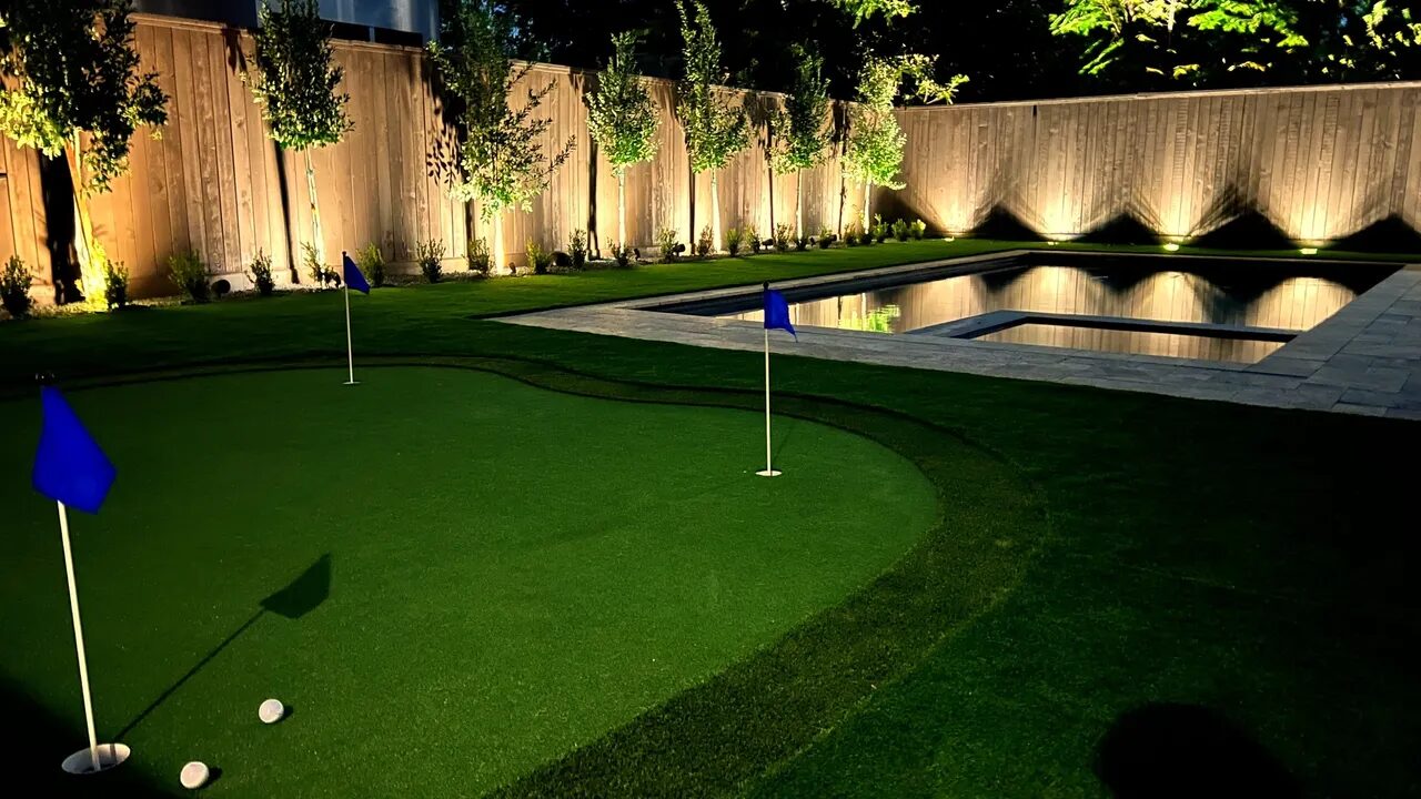 Putting green and pool in illuminated backyard.