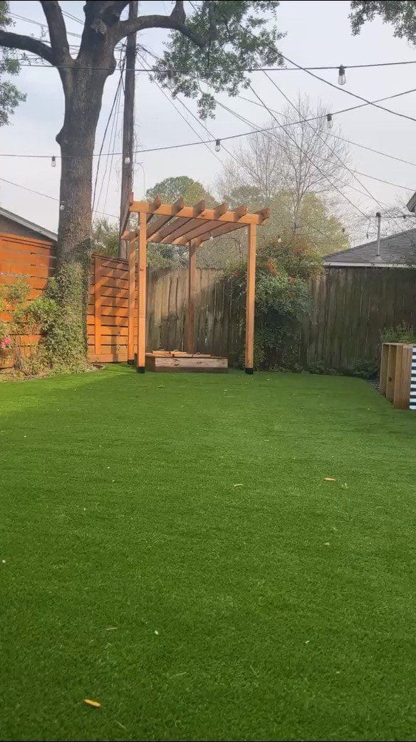 A backyard with green artificial grass and a wooden pergola near a brick wall.